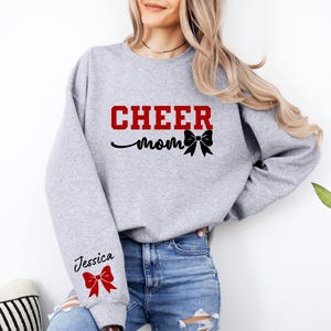 May include: Gray sweatshirt with the text "CHEER mom" in red and black with a black bow. The left sleeve has the text "Jessica" in black with a red bow.