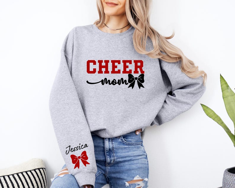 May include: Gray sweatshirt with the text "CHEER mom" in red and black with a black bow. The left sleeve has the text "Jessica" in black with a red bow.
