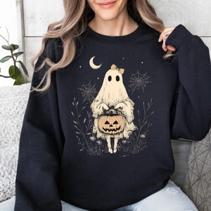 May include: Black sweatshirt with a white ghost holding a jack-o'-lantern. The ghost has a pink bow and is surrounded by spiderwebs and stars.