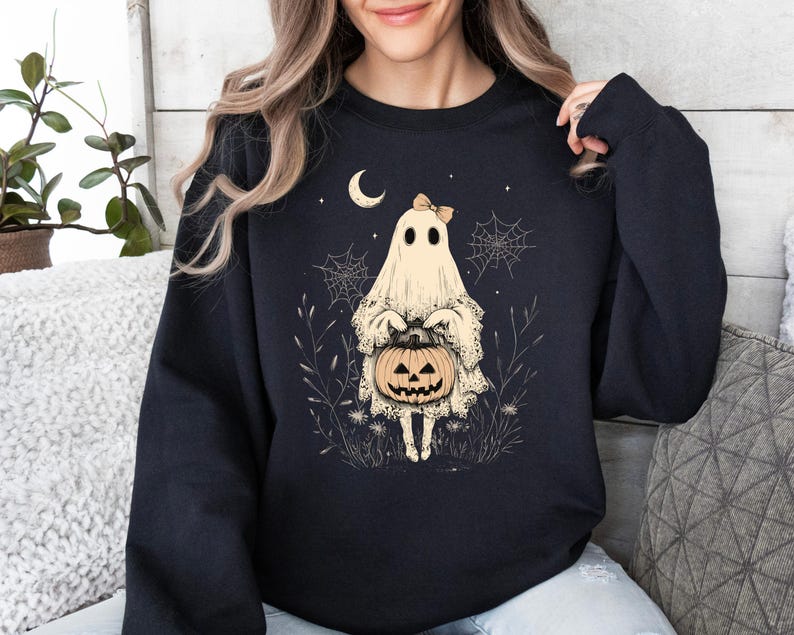 May include: Black sweatshirt with a white ghost holding a jack-o'-lantern. The ghost has a pink bow and is surrounded by spiderwebs and stars.