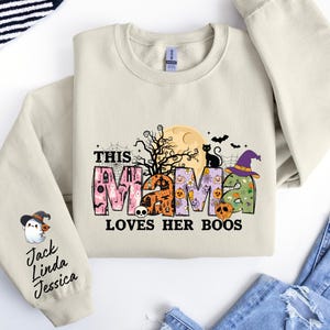 May include: A beige sweatshirt with a graphic print of the word "mama" decorated with Halloween-themed elements like pumpkins, bats, and a ghost. The text "This mama loves her boos" is printed below the graphic. The sleeve has a ghost graphic and the text "Jack Linda Jessica" printed on it.