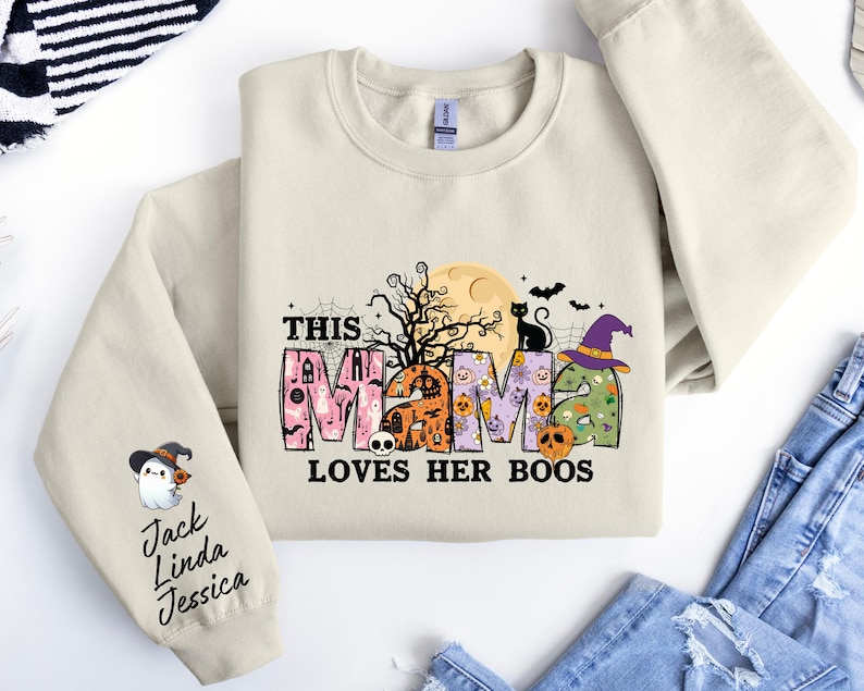 May include: A beige sweatshirt with a graphic print of the word "mama" decorated with Halloween-themed elements like pumpkins, bats, and a ghost. The text "This mama loves her boos" is printed below the graphic. The sleeve has a ghost graphic and the text "Jack Linda Jessica" printed on it.