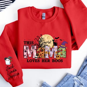 May include: Red sweatshirt with a Halloween-themed design that says "This Mama Loves Her Boos". The design features a black cat, a witch's hat, and a full moon. The sleeve has a ghost with a witch's hat and the names Jack, Linda, and Jessica.