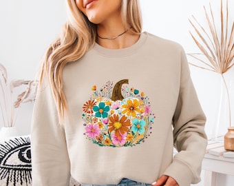 May include: Beige sweatshirt featuring a colorful floral pumpkin design. The pumpkin is filled with orange, pink, teal, and yellow flowers, with a brown stem. The design is centered on the front of the sweatshirt.