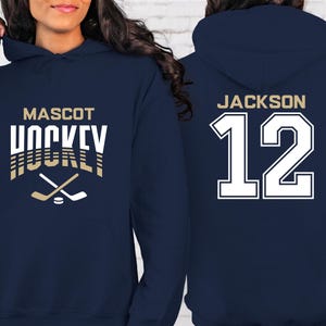 K&ouml;nnte beinhalten: Zwei marineblaue Kapuzenpullover. Ein Pullover hat den Text "MASCOT HOCKEY" in Wei&szlig; mit goldenen Akzenten und eine Grafik von zwei gekreuzten Hockeyschl&auml;gern. Der andere Pullover hat den Text "JACKSON 12" in Wei&szlig; mit goldener Umrandung.