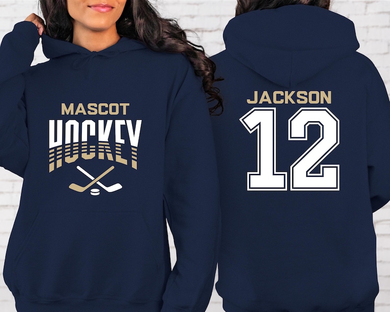 K&ouml;nnte beinhalten: Zwei marineblaue Kapuzenpullover. Ein Pullover hat den Text "MASCOT HOCKEY" in Wei&szlig; mit goldenen Akzenten und eine Grafik von zwei gekreuzten Hockeyschl&auml;gern. Der andere Pullover hat den Text "JACKSON 12" in Wei&szlig; mit goldener Umrandung.