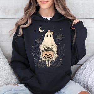 May include: Black hoodie with a white ghost holding a jack-o'-lantern. The ghost has a pink bow and is surrounded by spiderwebs and leaves.