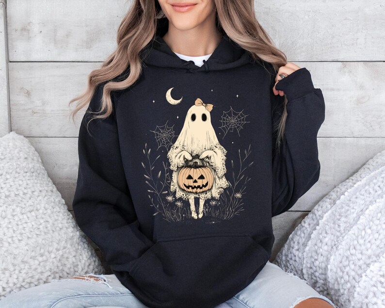 May include: Black hoodie with a white ghost holding a jack-o'-lantern. The ghost has a pink bow and is surrounded by spiderwebs and leaves.