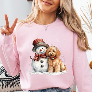 May include: A light pink sweatshirt featuring a watercolor illustration of a snowman wearing a hat and scarf, and a brown dog. The snowman has a carrot nose and button eyes. The dog is sitting next to the snowman.