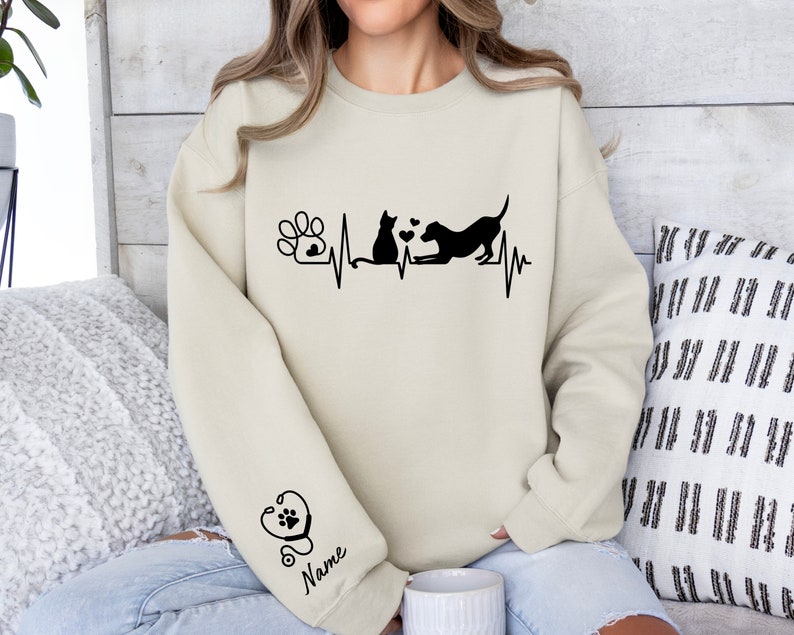 May include: A beige sweatshirt with a black paw print, cat, dog, and heartbeat graphic. The text "Name" is on the sleeve with a stethoscope graphic.