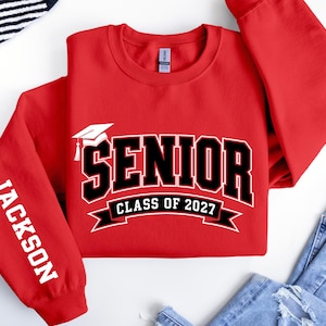 May include: A red long-sleeve sweatshirt featuring a white graduation cap icon and the text SENIOR CLASS OF 2027 in black with white outlines. The left sleeve displays the name JACKSON in white capital letters printed vertically.