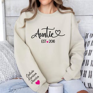 May include: A beige sweatshirt with the text "Auntie EST 2016" and a pink heart. The sleeve has the names "Calvin, Karissa, Jessica" and a pink heart.