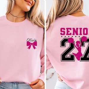 May include: A light pink crewneck sweatshirt featuring a small black script name Olivia and a hot pink bow on the front left chest. The back displays the word SENIOR in hot pink above the large black numbers 27 with a hot pink cheerleader graphic.