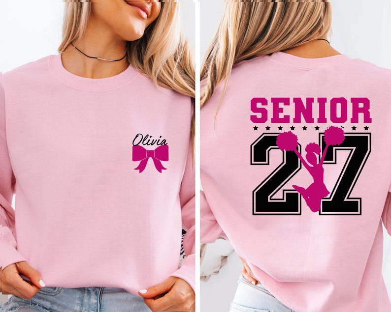 May include: A light pink crewneck sweatshirt featuring a small black script name Olivia and a hot pink bow on the front left chest. The back displays the word SENIOR in hot pink above the large black numbers 27 with a hot pink cheerleader graphic.