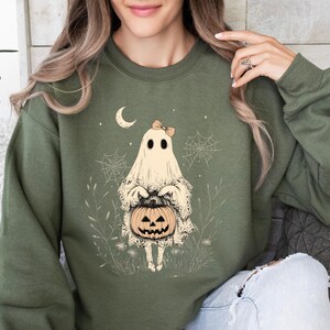May include: A green sweatshirt with a graphic of a ghost holding a jack-o'-lantern. The ghost is wearing a bow and has a friendly expression. The graphic is surrounded by a whimsical design of spiderwebs and leaves.