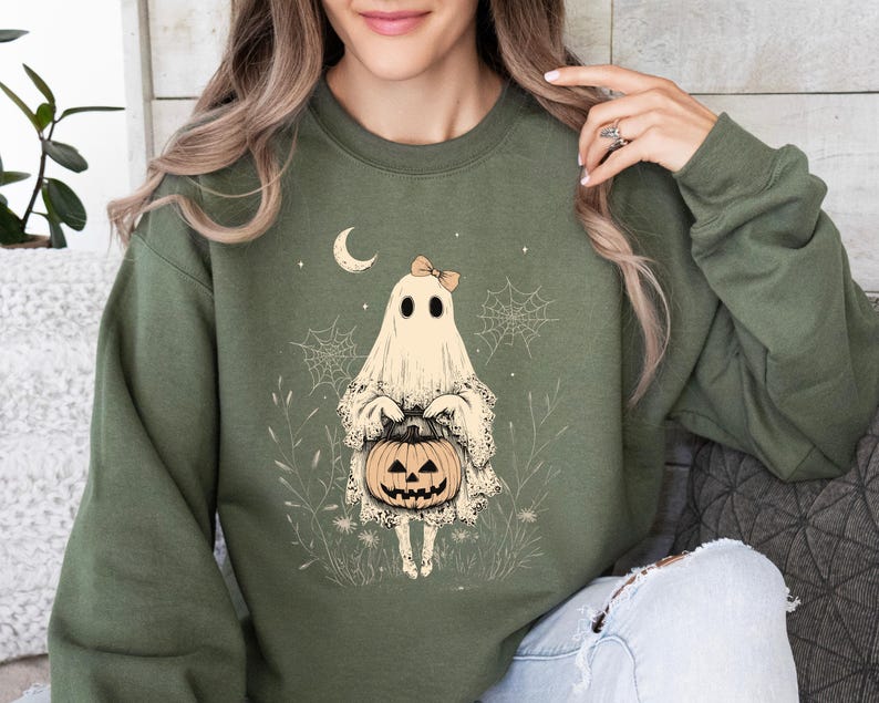 May include: A green sweatshirt with a graphic of a ghost holding a jack-o'-lantern. The ghost is wearing a bow and has a friendly expression. The graphic is surrounded by a whimsical design of spiderwebs and leaves.