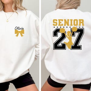 May include: A white crewneck sweatshirt featuring a gold bow graphic and the name Olivia on the front left chest. The back displays the text SENIOR in gold, the number 27 in black with white outlines, and a gold silhouette of a cheerleader.