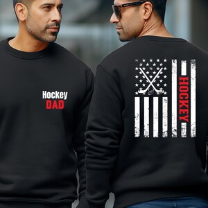 May include: Black sweatshirt with a white American flag design and the words "Hockey Dad" in red and white on the front. The back of the sweatshirt has a white American flag design with hockey sticks and a puck in the centre and the word "Hockey" in red on the right side.