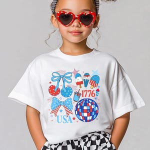 Puede incluir: Una camiseta blanca con un dise&ntilde;o patri&oacute;tico en rojo, blanco y azul. El gr&aacute;fico incluye lazos, polos de hielo, una bola de discoteca y los textos "USA" y "1776". La persona lleva gafas de sol en forma de coraz&oacute;n.