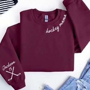 May include: A maroon sweatshirt with the words "hockey mama" in white script on the chest. The left sleeve features the name "Jackson" and the number "15" with crossed hockey sticks. The sweatshirt is folded.