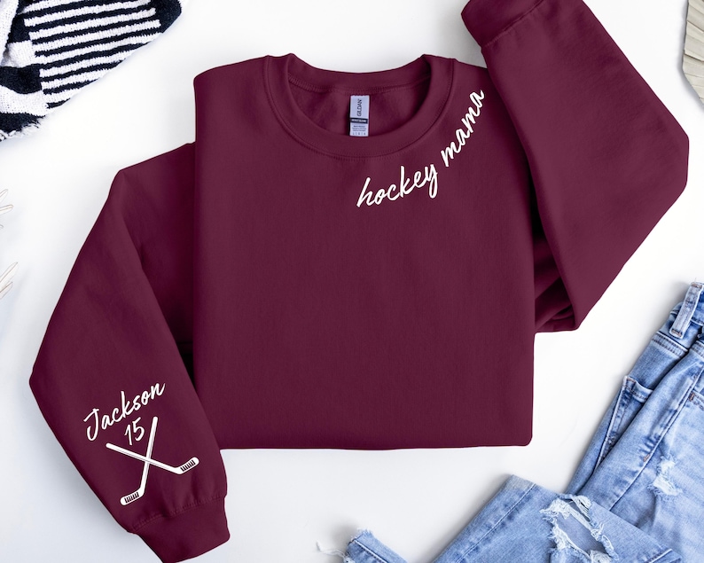 May include: A maroon sweatshirt with the words "hockey mama" in white script on the chest. The left sleeve features the name "Jackson" and the number "15" with crossed hockey sticks. The sweatshirt is folded.