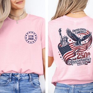 Pode incluir: Camiseta rosa claro com um design circular na frente com o texto "1776 2026". A parte de tr&aacute;s apresenta um design patri&oacute;tico com a Est&aacute;tua da Liberdade, uma &aacute;guia e a bandeira americana, com o texto "United States of America 250th Anniversary 1776-2026".