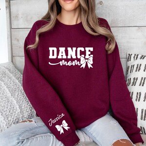 Pu&ograve; includere: Felpa color borgogna con il testo "Dance Mom" e un fiocco bianco. La felpa ha un fiocco bianco sulla manica con il nome "Jessica" scritto sopra.