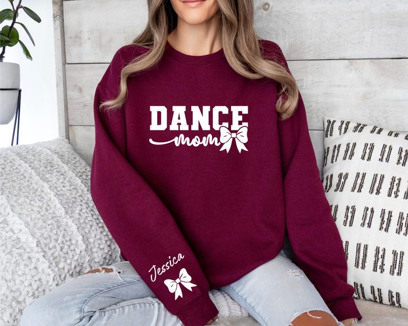 Pu&ograve; includere: Felpa color borgogna con il testo "Dance Mom" e un fiocco bianco. La felpa ha un fiocco bianco sulla manica con il nome "Jessica" scritto sopra.