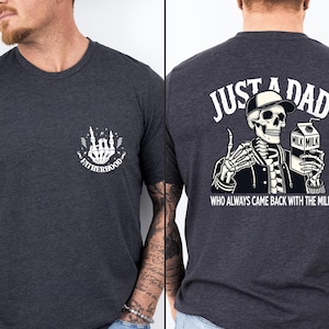 May include: Dark gray t-shirt with a white graphic on the front that says "FATHERHOOD" with a skeleton hand design. The back of the shirt features a skeleton wearing a hat and jacket, holding a milk carton, with the text "JUST A DAD WHO ALWAYS CAME BACK WITH THE MILK."