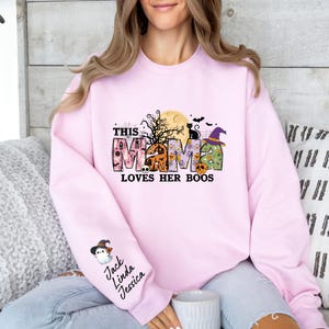 May include: Pink sweatshirt with a Halloween-themed graphic that says "This Mama Loves Her Boos". The graphic features a witch, a cat, and a house. The sweatshirt also has a patch on the sleeve with the names "Jack, Linda, Jessica".