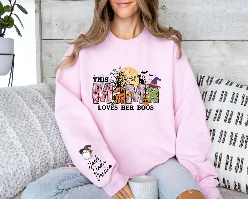 May include: Pink sweatshirt with a Halloween-themed graphic that says "This Mama Loves Her Boos". The graphic features a witch, a cat, and a house. The sweatshirt also has a patch on the sleeve with the names "Jack, Linda, Jessica".