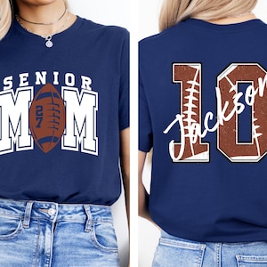 May include: A navy blue t-shirt featuring a front graphic with the text SENIOR MOM and a brown football with the number 27. The back graphic displays the number 10 in a brown football texture with the name Jackson written in white script across it.