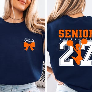 May include: A navy blue crewneck sweatshirt featuring a small orange bow graphic with the name Olivia in white script on the front left chest. The back displays the word SENIOR in orange above large white numbers 27 with a cheerleader graphic.