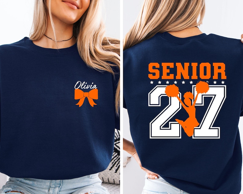 May include: A navy blue crewneck sweatshirt featuring a small orange bow graphic with the name Olivia in white script on the front left chest. The back displays the word SENIOR in orange above large white numbers 27 with a cheerleader graphic.