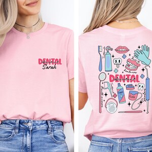 May include: Pink t-shirt with a dental assistant design on the back. The design includes a toothbrush, toothpaste, floss, mouth mirror, dental tools, and a pair of dentures. The front of the shirt says "Dental Assistant Sarah".
