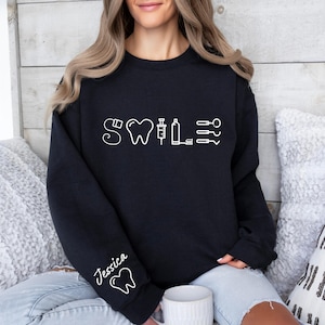 May include: Black sweatshirt with a white graphic of a tooth, a syringe, and dental tools. The text "Smile" is above the graphic. The sweatshirt has the name "Jessica" written on the sleeve.