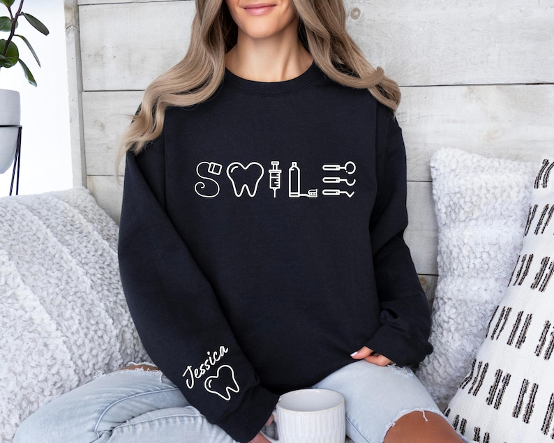 May include: Black sweatshirt with a white graphic of a tooth, a syringe, and dental tools. The text "Smile" is above the graphic. The sweatshirt has the name "Jessica" written on the sleeve.
