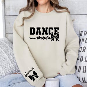 Pu&ograve; includere: Una felpa color crema con il testo "DANCE mom" e un fiocco nero. La felpa ha un fiocco nero sulla manica sinistra con il nome "Jessica" scritto sopra.