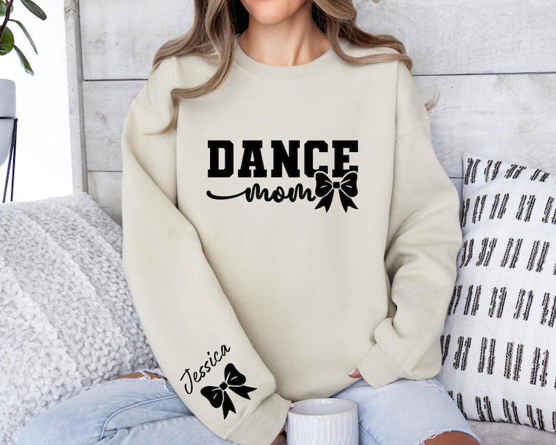 Pu&ograve; includere: Una felpa color crema con il testo "DANCE mom" e un fiocco nero. La felpa ha un fiocco nero sulla manica sinistra con il nome "Jessica" scritto sopra.
