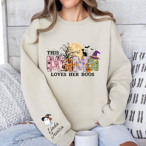 May include: A cream colored sweatshirt with a Halloween themed graphic that says "This Mama Loves Her Boos". The graphic features a black cat, a witch's hat, and a moon with a spooky tree.