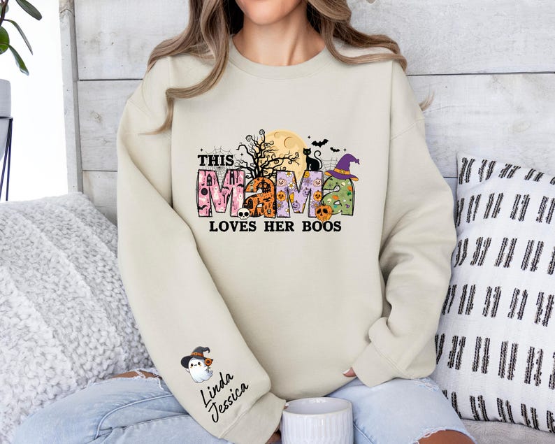 May include: A cream colored sweatshirt with a Halloween themed graphic that says "This Mama Loves Her Boos". The graphic features a black cat, a witch's hat, and a moon with a spooky tree.