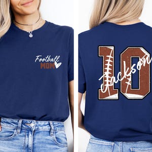 May include: Two navy blue t-shirts. The front of one shirt says "Football Mom" with a heart. The back of the other shirt has a large number 10 with the word "Jackson" written inside the number. The number is made to look like a football.