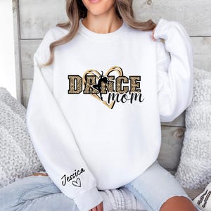 May include: White sweatshirt with a black and gold heart design that says "Dance Mom". The heart has a silhouette of a dancer inside. The sweatshirt has the name "Jessica" written on the sleeve with a small heart.