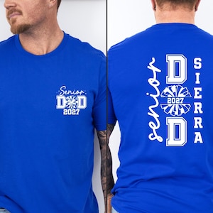 May include: A royal blue t-shirt featuring white graphic text. The front displays 'Senior DAD 2027' with a pom-pom icon. The back features the vertical text 'Senior DAD SIERRA' with a large pom-pom graphic and the year '2027' centered within the design.