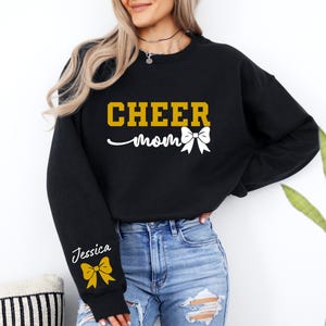 May include: Black sweatshirt with the text "CHEER mom" in gold letters and a white bow. The sleeve has the name "Jessica" in white letters and a gold bow.