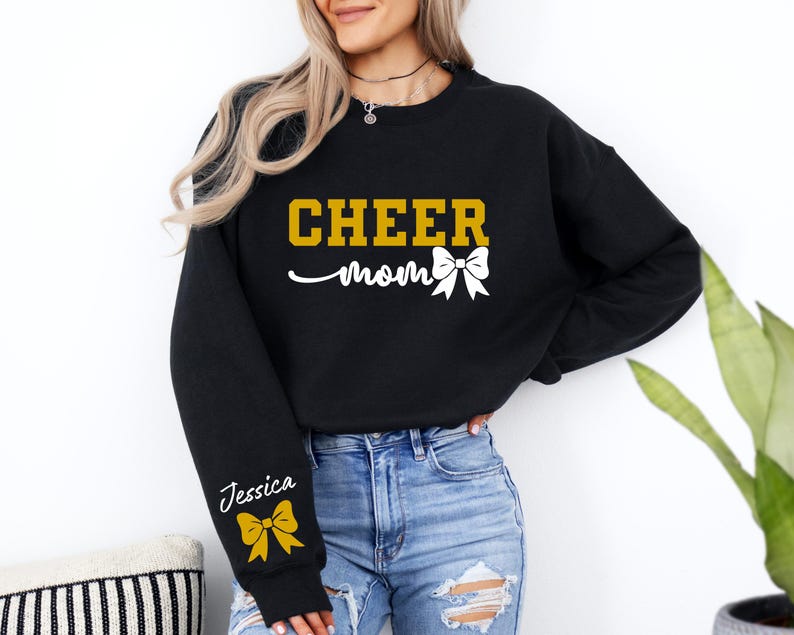 May include: Black sweatshirt with the text "CHEER mom" in gold letters and a white bow. The sleeve has the name "Jessica" in white letters and a gold bow.