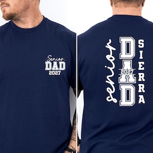May include: A navy blue short-sleeved t-shirt featuring white screen-printed text. The front displays 'Senior DAD 2027' in a small chest graphic. The back features a large vertical design with the words 'Senior', 'DAD', '2027', and 'SIERRA'.
