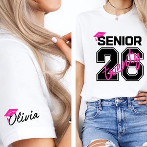 May include: White t-shirt with a black and pink graphic design. The design includes the word "SENIOR" above the number "20" in a black outlined font, with the word "Twenty" in pink. A pink graduation cap is also included. The sleeve has a pink graduation cap and the name "Olivia".
