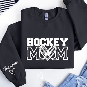 Pu&ograve; includere: Felpa nera con testo bianco che recita "Hockey Mom" con un cuore e bastoni da hockey. La manica sinistra ha un testo bianco che recita "Jackson" con un cuore.