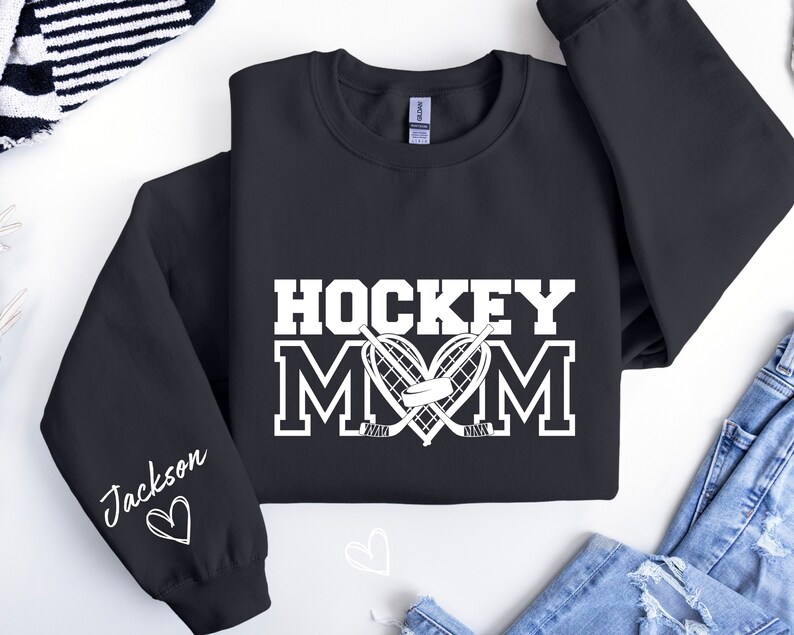 Pu&ograve; includere: Felpa nera con testo bianco che recita "Hockey Mom" con un cuore e bastoni da hockey. La manica sinistra ha un testo bianco che recita "Jackson" con un cuore.